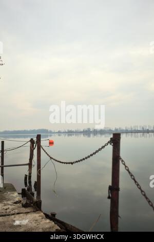 Chaîne de fer sur un coin par le bord d'une jetée au bord du lac, avec des bouées flottantes à proximité et un ciel nuageux coulé dans l'eau, dans le pays italien Banque D'Images