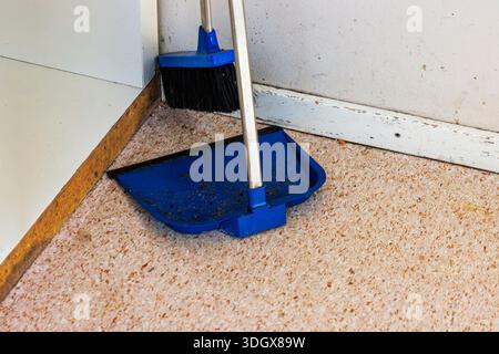 Gros plan d'une pelle à poussière bleue et d'un balai debout dans un coin sur un sol intérieur sale avec des débris visibles. Banque D'Images