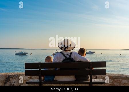 Mère dans un chapeau de soleil embrassant deux enfants sur un banc en bois regardant des yachts dans la mer calme pendant l'heure dorée du coucher du soleil. Concept de lien familial, holid côtier Banque D'Images