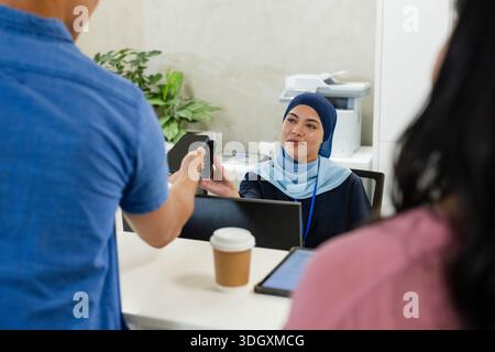 Réceptionniste féminine et visiteurs échangeant leur appareil de paiement à la réception, portant un hijab bleu Banque D'Images
