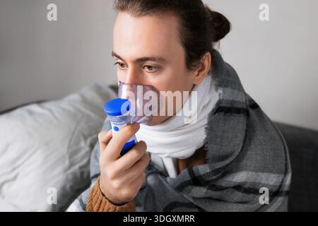 Un homme malade enveloppé dans une couverture utilise un inhalateur nébuliseur à mailles portatif assis sur un canapé, entouré de médicaments et de tissus pendant le répi à la maison Banque D'Images