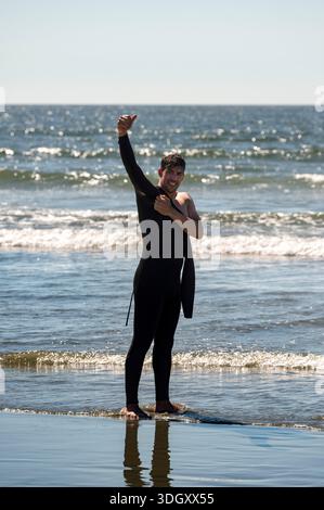 Un homme enfilant sa combinaison avec ses pieds dans l'eau prêt à surfer quelques vagues tueuses de l'océan à Tofino Canada Banque D'Images