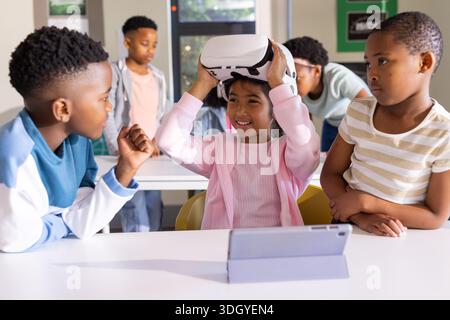 Divers enfants se réunissant en classe, regardant une fille asiatique tenant un casque VR blanc près de la tablette Banque D'Images