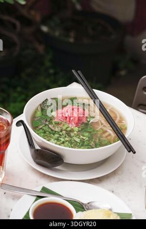 Un délicieux bol de soupe traditionnelle vietnamienne aux nouilles pho est magnifiquement présenté sur une table blanche. Il présente du bœuf rare tendre, des herbes vertes fraîches, Banque D'Images