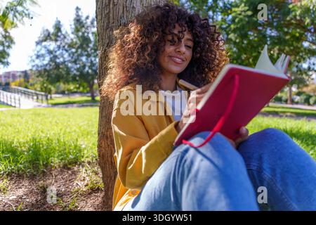 Femme aux cheveux bouclés souriant, écrivant ou journalisant à l'extérieur par une journée ensoleillée. Saisir des pensées et des idées dans un journal personnel Banque D'Images