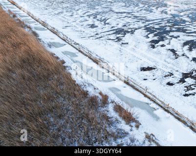 Paysage hivernal aérien d'un sentier côtier gelé et d'un marais enneigé en Estonie avec une clôture en bois et de grands roseaux. Banque D'Images