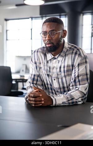 Homme afro-américain assis à la table de conférence dans un bureau moderne, portant des lunettes et une chemise à carreaux Banque D'Images