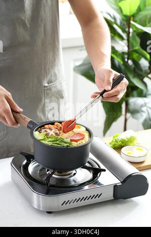 Personne cuisinant des nouilles instantanées avec des crevettes, des œufs et des légumes dans une petite casserole sur une cuisinière à gaz Banque D'Images