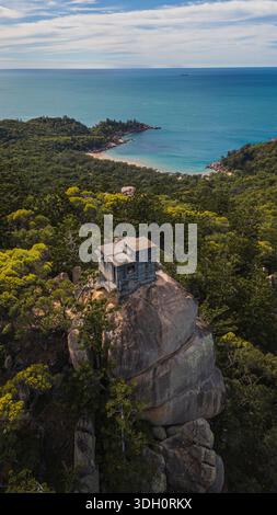 Vue aérienne d'un petit bâtiment carré perché au sommet d'un massif rocher altéré surplombant la mer Azur, entouré d'une forêt tropicale dense et verdoyante, Banque D'Images