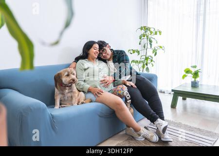 Femme enceinte heureuse et mari avec jambe prothétique se relaxant sur le canapé avec leur chien, profitant d'un moment paisible à la maison Banque D'Images