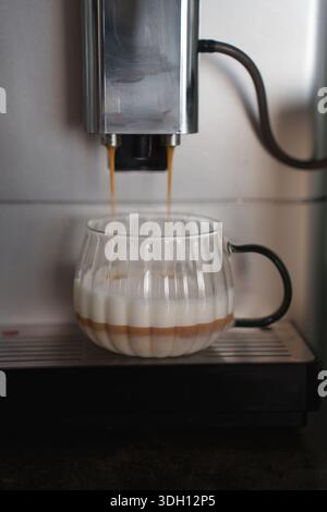 vue rapprochée d'une machine à espresso versant des couches de café et de lait dans une tasse à côtes transparente, mettant en valeur le savoir-faire d'un barista dans un ensemble de cuisine moderne Banque D'Images