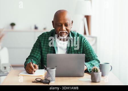 Homme senior utilisant un ordinateur portable à un bureau tout en prenant des notes dans une salle lumineuse Banque D'Images
