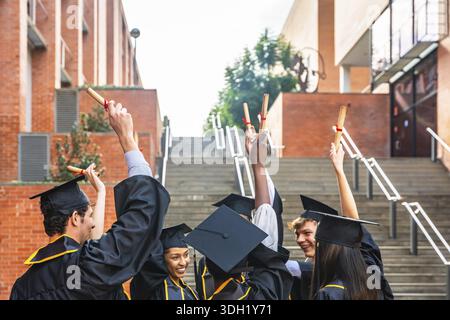 Groupe d'étudiants diplômés divers portant des casquettes et des robes académiques, élevant heureusement leurs rouleaux de diplôme dans la célébration sur un campus universitaire afte Banque D'Images