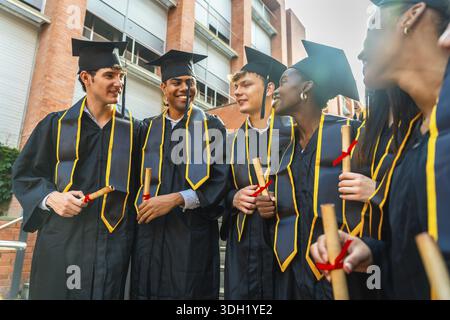Groupe d'étudiants diplômés divers portant des robes académiques et des casquettes, souriant tout en tenant leurs diplômes et célébrant à l'extérieur d'un bâtiment universitaire Banque D'Images