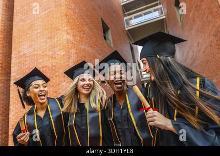 Groupe diversifié d'étudiantes heureuses célébrant leur diplôme universitaire, portant fièrement des casquettes et des robes tout en détenant des diplômes et exprimant J. Banque D'Images