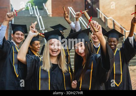 Groupe d'étudiants universitaires heureux et diversifiés obtenant leur diplôme, célébrant leur réussite scolaire ensemble, portant des casquettes et des robes de remise des diplômes, et fièrement Banque D'Images