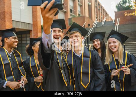 Groupe d'étudiants souriants divers diplômés de l'université, portant des casquettes et des robes académiques, détenant des diplômes, et enregistrant un moment partagé avec un Banque D'Images