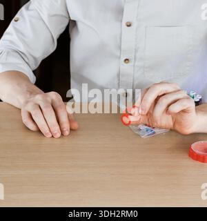 Gros plan de mains masculines tenant de minuscules ciseaux de broderie rouges au-dessus des aiguilles et des fils sur la table en bois Banque D'Images