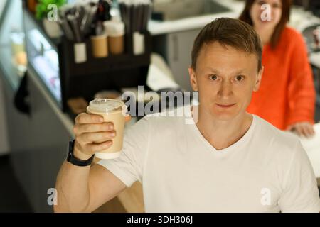 Un client masculin occasionnel soulève une tasse de café crémeuse vers la caméra dans un cadre de café lumineux avec le serveur souriant derrière Banque D'Images