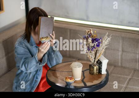 Jeune brune tient ludique muffin en forme de coeur près du visage tout en appréciant livre et café dans un café moderne confortable Banque D'Images