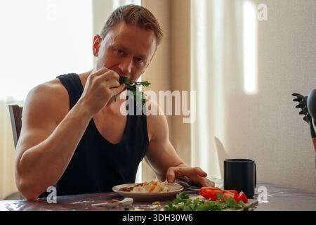 Masculin athlétique dans le débardeur consomme de l'herbe avec une fourchette dans la salade, des légumes dispersés sur la table à la maison. Banque D'Images