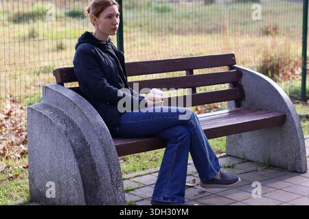 Angle latéral de femelle assise dans le parc d'automne, tenant la tasse à café, portant un Jean et une veste. Banque D'Images