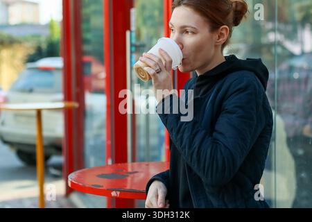 Jeune femme aux cheveux rouges dans une veste décontractée boit de tasse à emporter tout en se penchant sur la table à l'abri de bus à cadre rouge en ville Banque D'Images