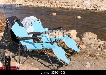 Deux chaises pliantes turquoise avec des vêtements dispersés et des objets près d'une rivière qui coule sur un rivage de galets dans un cadre naturel Banque D'Images