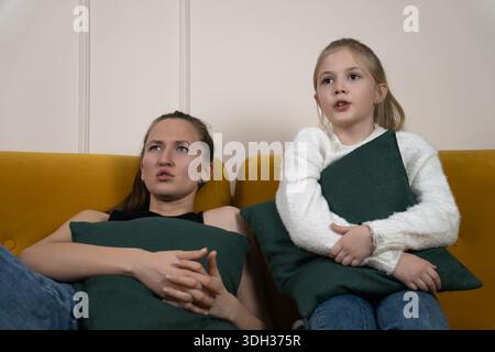 Mère et fille sur un canapé jaune avec des oreillers verts regarder un film effrayant ensemble, se serrant et montrant la peur, le suspense et la famille proche Banque D'Images