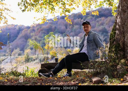 L'homme pointe vers le ciel avec une main, l'autre sur le clavier de l'ordinateur portable, assis sur le journal avec le café, le feuillage d'automne et les montagnes lointaines en vue. Banque D'Images