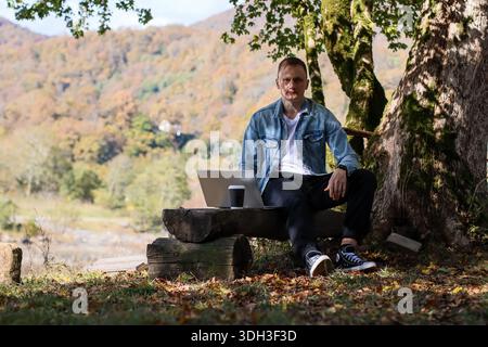 Homme détendu assis avec ordinateur portable ouvert et café sur banc en bois, feuilles d'automne et montagnes lointaines dans la scène forestière tranquille Banque D'Images