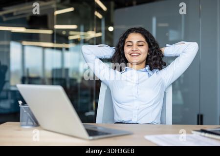 Jeune femme d'affaires indienne souriant les yeux fermés, assise dans une chaise de bureau et étirant derrière sa tête, profitant d'une pause détendue et satisfaite après avoir terminé le travail Banque D'Images
