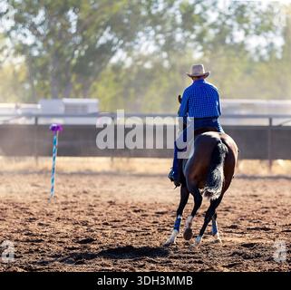 Un cavalier vêtu d'une chemise à carreaux bleue guida son cheval à travers une arène poussiéreuse vers un poteau de marquage rayé bleu et blanc. Banque D'Images