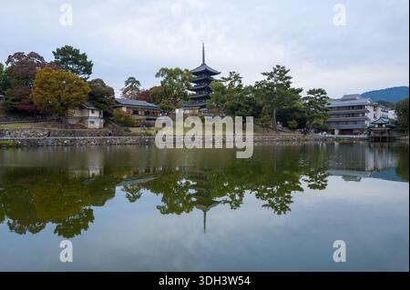 Nara, Japon - 4 novembre 2023 : L'image montre la pagode de cinq étages du temple Kofuku-ji et les bâtiments environnants reflétés dans l'étang de Sarusawa, avec des arbres et des gens visibles le long du front de mer sous un ciel nuageux. Banque D'Images