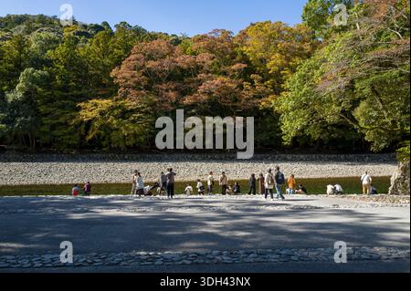 ISE, Japon - 29 octobre 2023 : des groupes de personnes se tiennent debout et s'assoient le long du bassin de purification de Mitarashiba près de la rivière, avec des arbres montrant le feuillage d'automne en arrière-plan. Banque D'Images