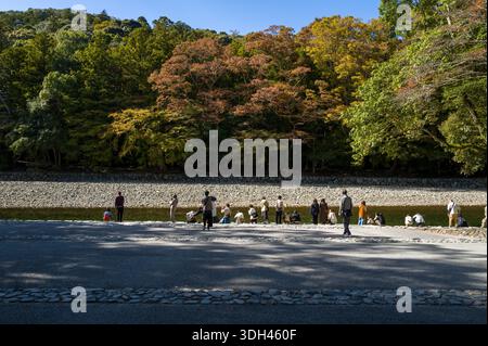 ISE, Japon - 29 octobre 2023 : des groupes de personnes se tiennent debout et s'assoient le long du bassin de purification de Mitarashiba près de la rivière, avec des arbres montrant le feuillage d'automne en arrière-plan. Banque D'Images