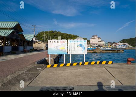 Nachikatsuura, Japon - 26 octobre 2023 : deux panneaux illustrés avec du texte japonais, y compris un avertissement sur le danger de la mer et un message sur la propreté du port, sont affichés au bord d'un quai avec de l'eau, des bâtiments et des bateaux visibles en arrière-plan. Banque D'Images