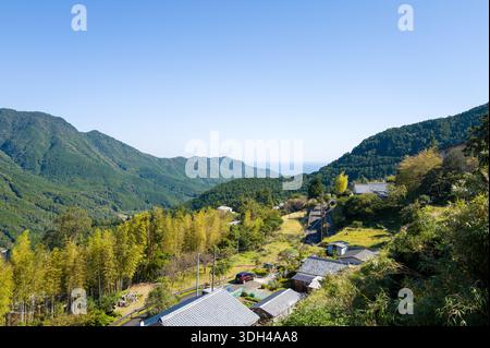 Nachisan, Japon - 26 octobre 2023 : des maisons japonaises traditionnelles aux toits de tuiles sont vues dans un petit village de montagne, avec des collines boisées et une vue sur la mer au loin sous un ciel clair. Banque D'Images