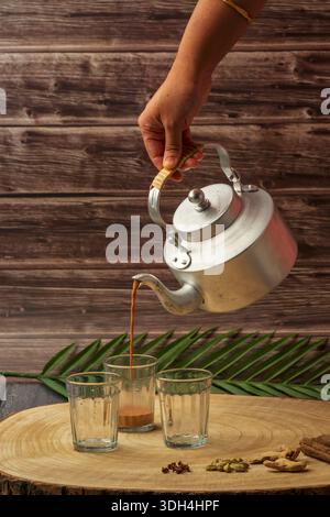 Une personne verse du thé masala dans une tasse de chai de trois verres. Les tasses sont sur une table en bois. Coupe chai versé hors de la bouilloire à thé. Banque D'Images