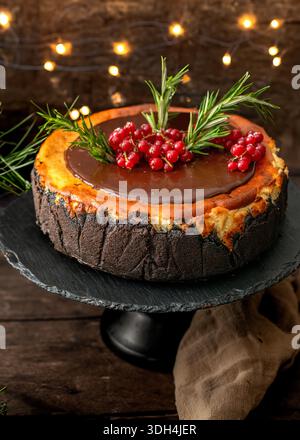Gros plan d'un cheesecake basque rustique brûlé sur un stand noir, garni d'une ganache au chocolat lisse, de groseilles rouges fraîches et de brins de romarin. Banque D'Images