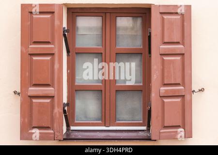Une seule fenêtre encadrée par des volets en bois rouge-brun montés sur un mur extérieur beige. Les vitres fermées et les charnières altérées transmettent le charme rustique et le temps Banque D'Images