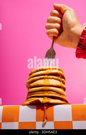 Une scène ludique de petit-déjeuner avec une pile de crêpes arrosées de sirop maintenues par une fourchette, sur fond rose vif avec une nappe à carreaux Banque D'Images
