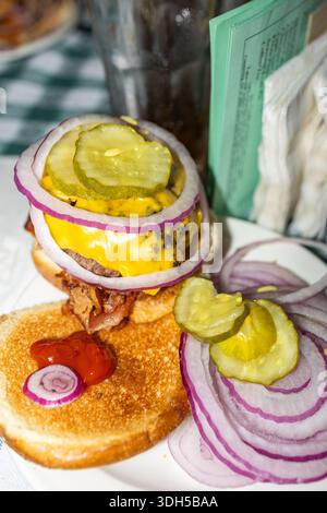 Un délicieux cheeseburger garni d'oignons rouges, de cornichons et de fromage fondu est ouvert sur un pain grillé avec une cuillère de ketchup. L'Amérique parfaite Banque D'Images