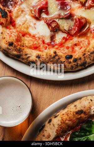 Gros plan de pizzas sur une assiette blanche, accompagnée d'une bière mousseuse. La scène capture l'essence d'une pizzeria confortable avec des tables en bois rustiques. Banque D'Images