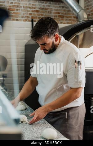 Un pizzeria qualifié fabrique de la pâte sur un comptoir en granit, vêtu d'une veste de chef blanche, entourée d'un décor rustique de briques et de TI Banque D'Images