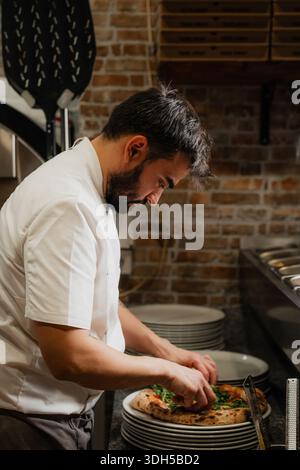 Un fabricant de pizza qualifié garnit une pizza napolitaine fraîchement cuite dans une pizzeria confortable. La chaleur de l'intérieur en brique complète le presentat artistique Banque D'Images