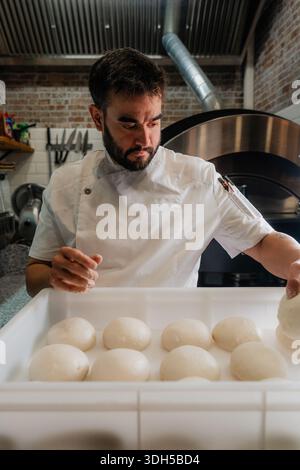 Un pizzeria qualifié inspecte les boules de pâte, assurant une texture et une consistance parfaites avant la cuisson. Le cadre est rustique, mettant en évidence Banque D'Images