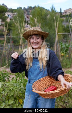 Une femme dans un chapeau de paille récolte joyeusement les légumes dans son jardin. Elle porte une combinaison en denim et tient un panier en osier avec des produits fraîchement cueillis, sur Banque D'Images