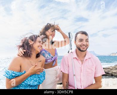 Un groupe diversifié d'amis, y compris une personne aveugle, profitant d'une journée ensoleillée à la plage. Leur interaction joyeuse incarne l’amitié et l’inclusion Banque D'Images