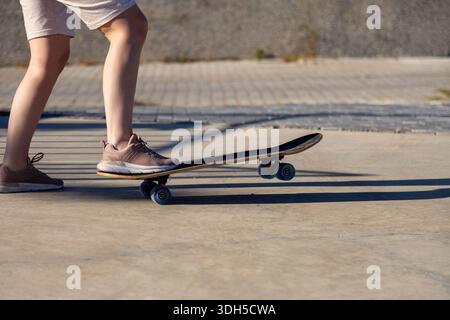 Un adolescent portant des shorts et des baskets manœuvre habilement une planche à roulettes sur un chemin en béton ensoleillé, projetant une longue ombre sur cette brillante journée d'été. Banque D'Images